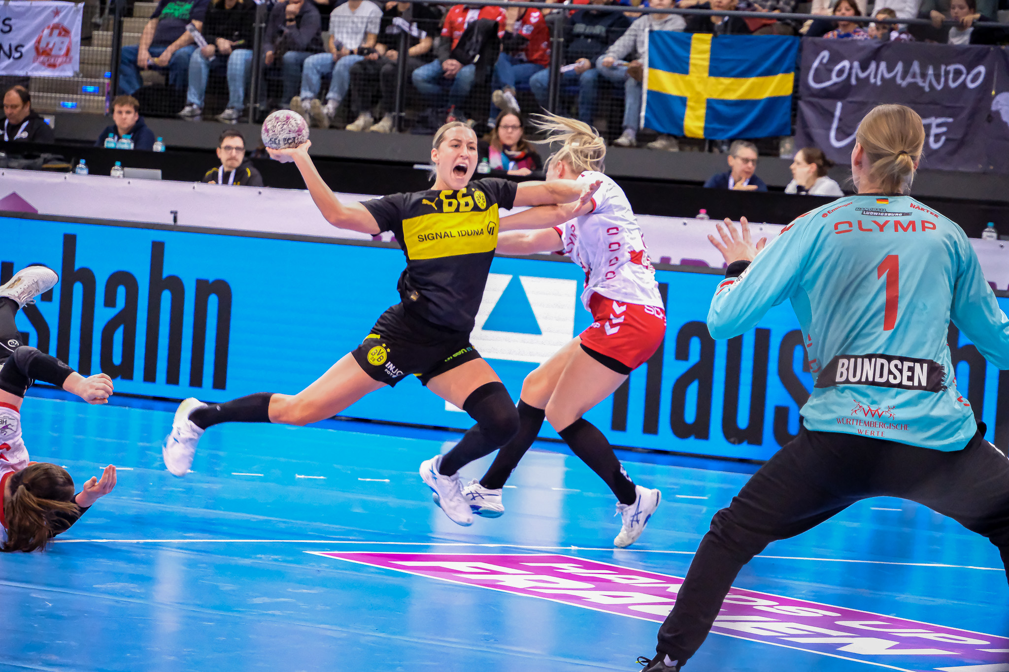 Eine Frauenhandballspielerin in einem schwarz-gelben Trikot springt, um den Ball zu schießen, während sie von einem Gegner in Weiß und Rot verteidigt wird. Der Torwart steht bereit, und die Zuschauer schauen von der Tribüne aus zu.
