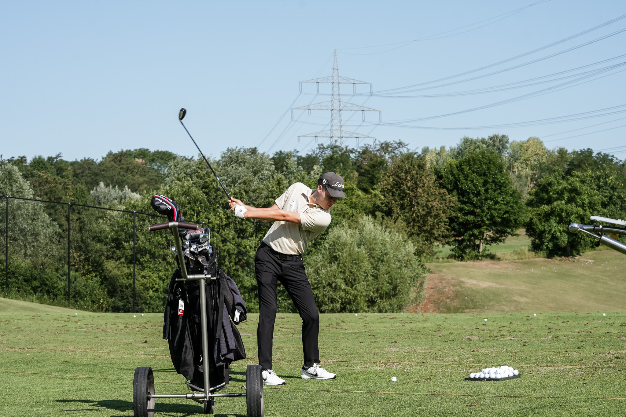 Ein Golfer in einem beigen Hemd und einer schwarzen Hose übt einen Schlag auf einer grasbewachsenen Driving Range, mit einer Golftasche und einem Schlägerwagen in der Nähe. Golfbälle liegen auf dem Boden verstreut, und im Hintergrund sind Bäume und Stromleitungen zu sehen.