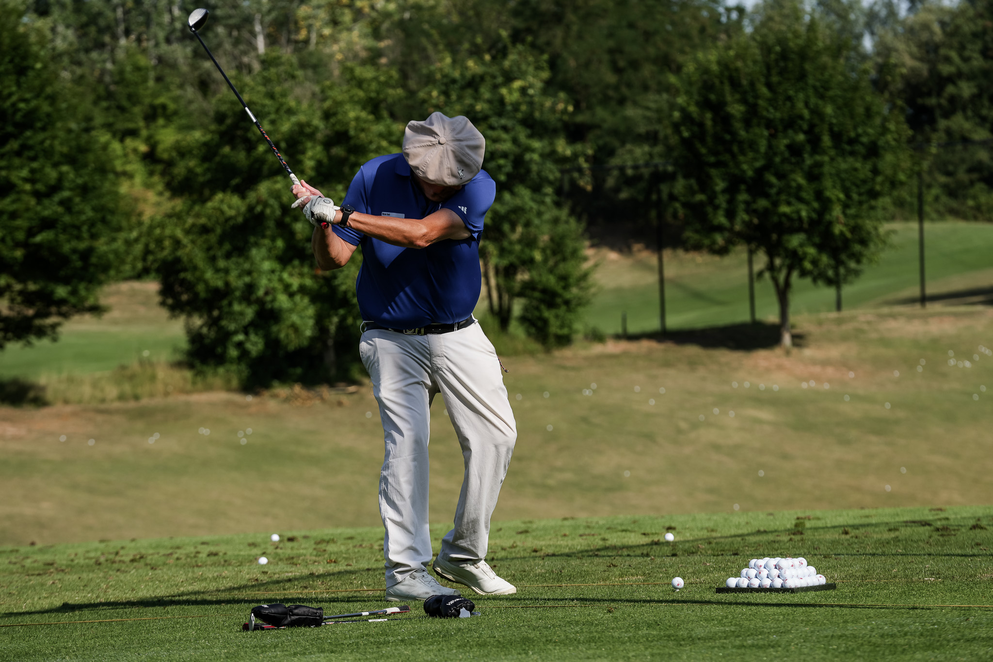 Ein Golfer in blauem Hemd und beiger Hose schwingt einen Schläger auf einer Driving Range, während in der Nähe ein Haufen Golfbälle auf dem Rasen liegt und Bäume im Hintergrund zu sehen sind.