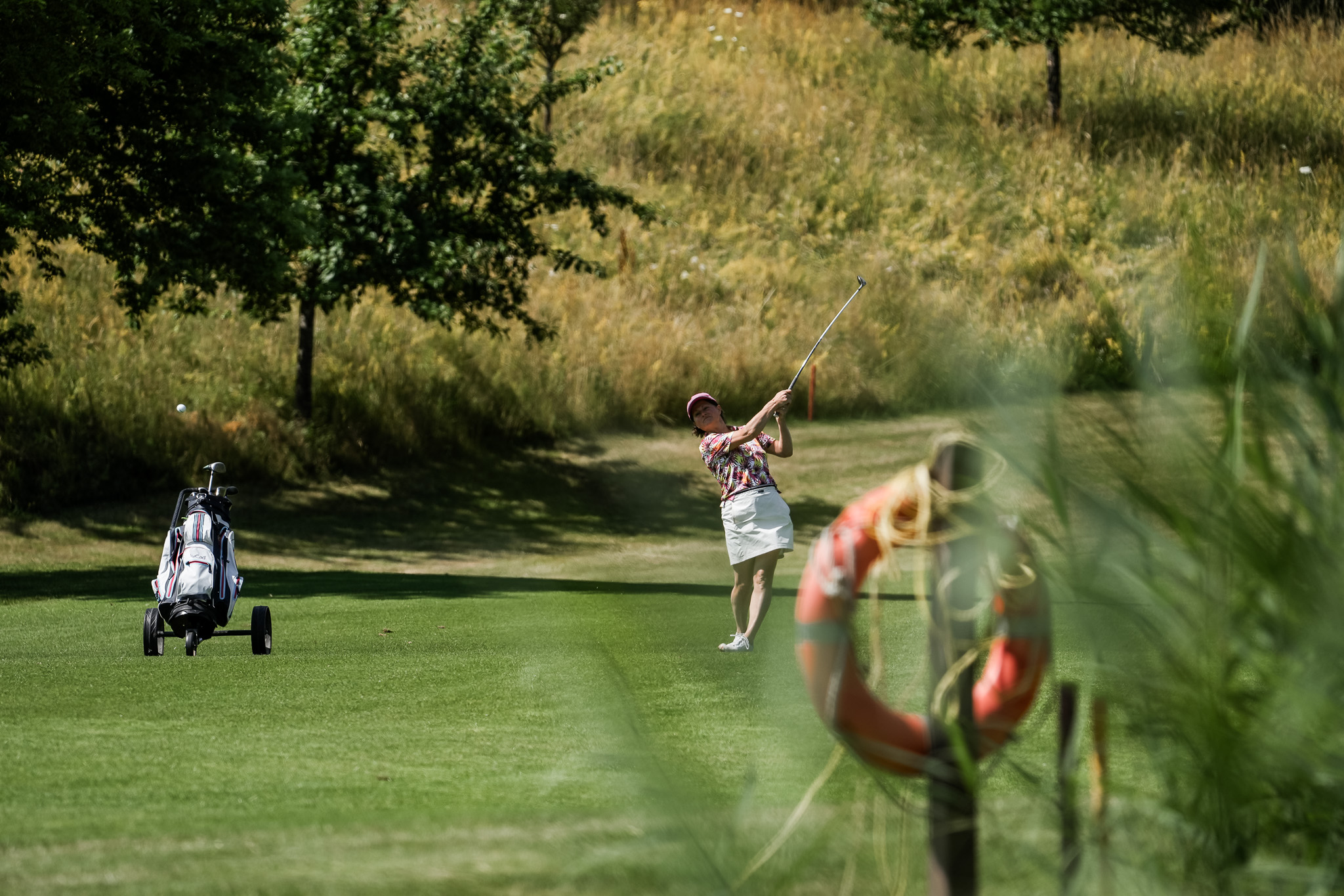 Eine Person, die einen Hut und ein geblümtes Hemd trägt, schwingt einen Golfschläger auf einem sonnigen Golfplatz, mit einer Golftasche auf einem Wagen in der Nähe und grünen Bäumen und Gras im Hintergrund. Im Vordergrund ist eine Rettungsweste zu sehen.