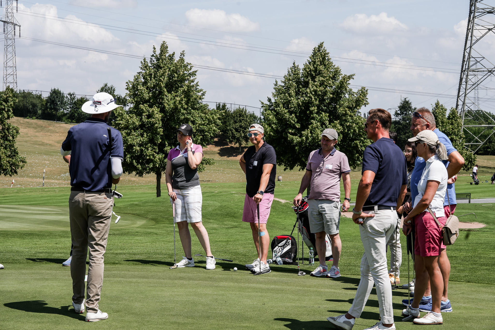 Eine Gruppe von Personen steht auf einem Golfplatz in der Nähe eines Putting Green und hört einem Golflehrer zu. Mehrere Golftaschen stehen in der Nähe, und die Szene ist sonnig mit Bäumen und Stromleitungen im Hintergrund.