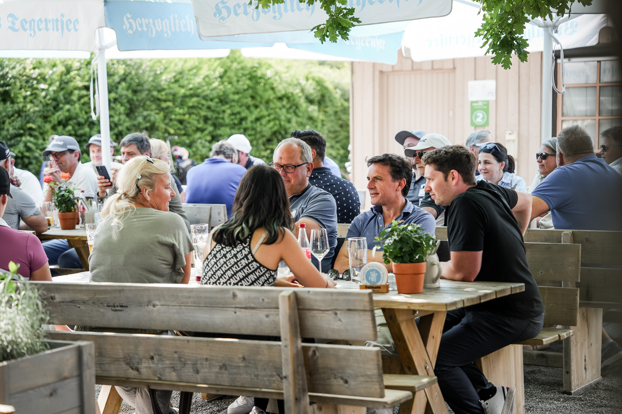 Eine Gruppe von Menschen sitzt an hölzernen Picknicktischen im Freien, unterhält sich und genießt Getränke unter großen Sonnenschirmen, während sich im Hintergrund weitere Menschen unterhalten.